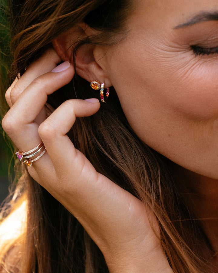Een vrouw met gesloten ogen en loshangend haar raakt haar oor aan, met aan haar vingers de Geelgouden Oorringen van Excellent Jewelry met Regenboog Saffier in Chatonzetting en gouden ringen bezet met veelkleurige saffieren.