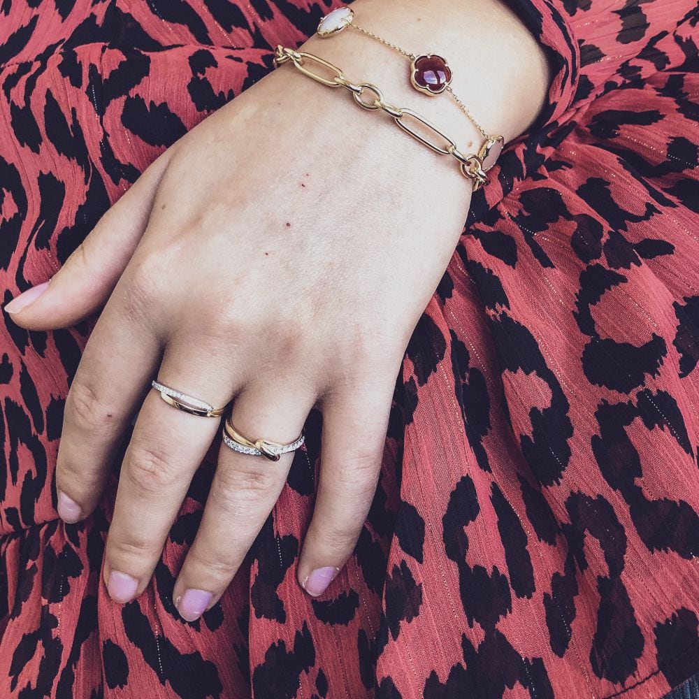 Een hand met lichtroze nagellak rust op een jurk met rode en zwarte dierenprint. De persoon draagt twee ringen, waaronder de Excellent Jewelry 14-Karaats Bicolor Gouden Damesring met Briljant Geslepen Diamanten, en twee gouden armbanden, waarvan één met een rode kralenbedel.