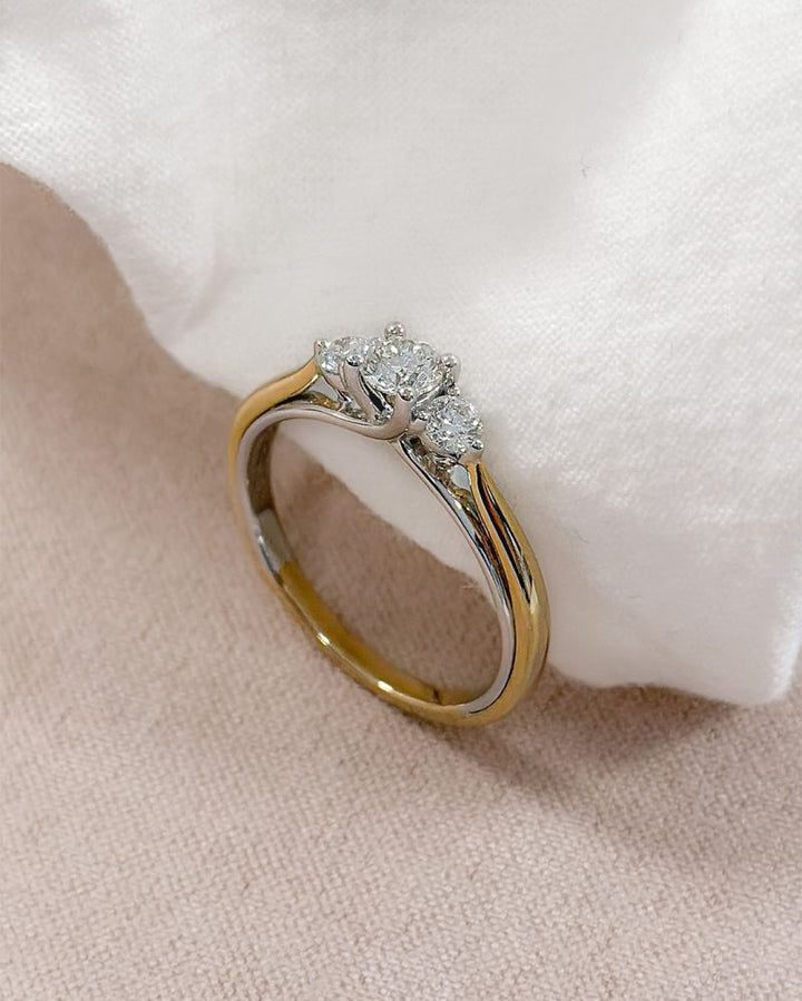 De Bicolor Geel- en Witgouden Damesring met Diamant (0.51 crt) van Excellent Jewelry, 14 karaat, schittert met drie ronde diamanten bovenop en ligt elegant op een zachte witte stof.