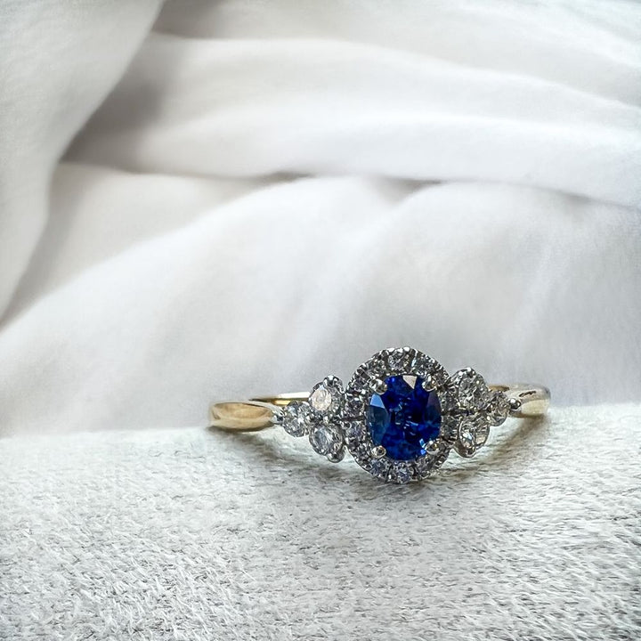 De 14-karaats bicolor gouden damesring met blauwe saffier en vintage diamanten zetting van Excellent Jewelry ligt op een zachte witte stof.