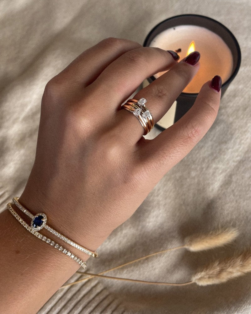 Een hand met bordeauxrode nagels draagt meerdere ringen, waaronder de 14k Bicolor Gold Solitaire Ring van Excellent Jewelry met een smaragdgeslepen diamant van 0,22ct, plus twee delicate armbanden - één met een blauwe steen die rust bij een brandende kaars op een beige structuurdecor.