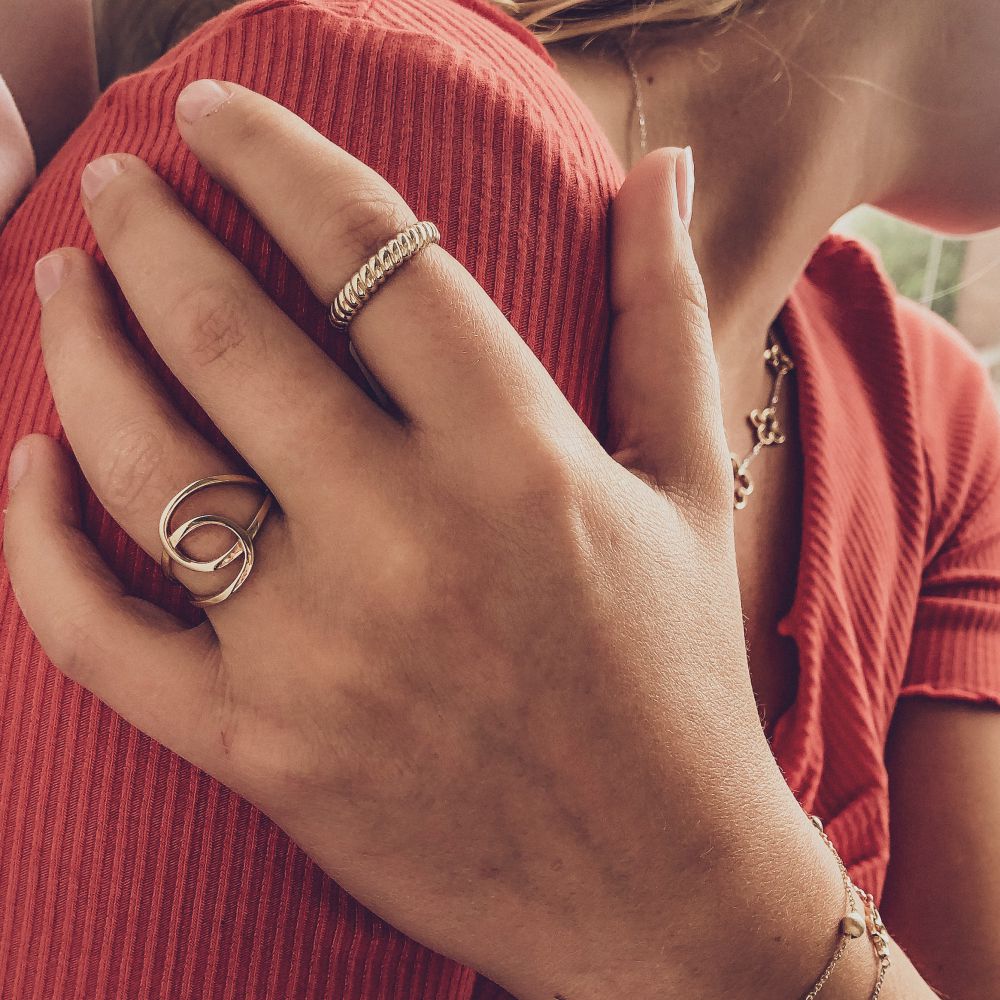 Een close-up toont een persoon in een rood geribbeld topje, hand op schouder die de Geelgouden Damesring - 14 Karaat Goud met Strak Design van Excellent Jewelry draagt, gecombineerd met andere gouden stukken. Hun nagels zijn schoon en ongepolijst.