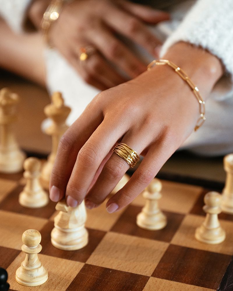 Een hand die de Speelse Geelgouden Damesring van Excellent Jewelry draagt, een 14-karaats gouden brede ring met speelse banden, beweegt een witte pion op een houten schaakbord met schaakstukken op de achtergrond.