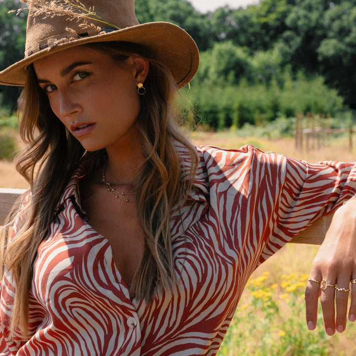 Een vrouw met een bruine hoed en rood-wit zebraprint shirt leunt op een houten hek onder de zon, draagt gouden sieraden waaronder de Excellent Jewelry Geelgouden Damesring met Speels Design - 14 Karaat Goud. Bomen en haar golvende haar maken het stralende tafereel compleet.