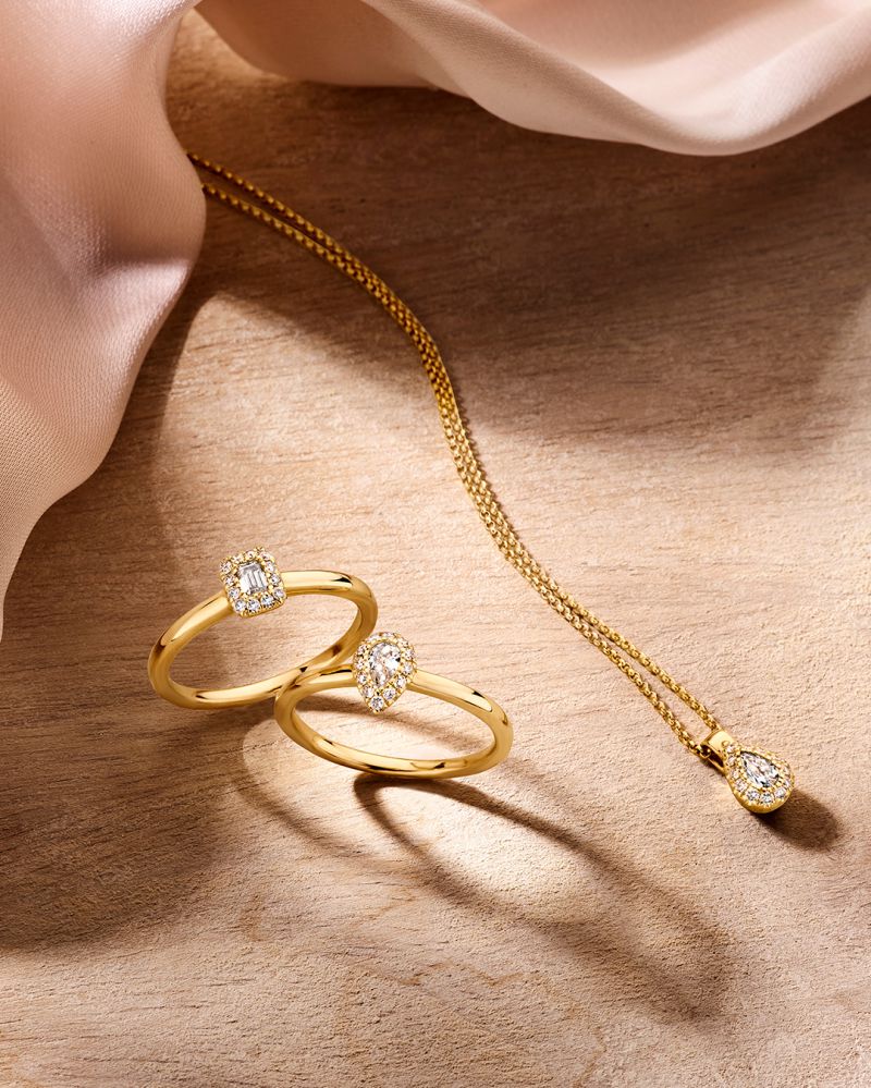 Twee verfijnde geelgouden ringen met baguette diamant 0.24 crt van Excellent Jewelry en een gouden ketting met peervormige diamant hanger, stijlvol gepresenteerd op hout naast zacht lichtroze stof.