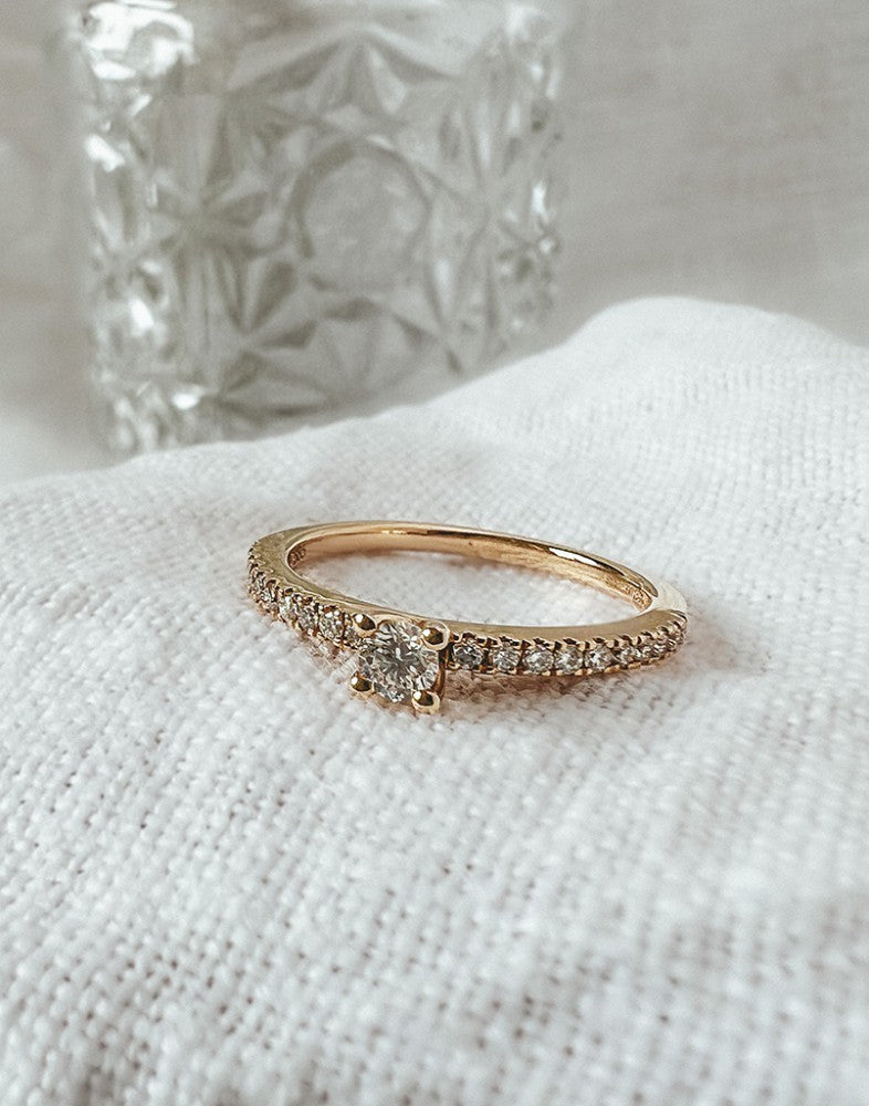 De Schitterende Geelgouden Solitair Ring met Zijstenen van Excellent Jewelry (14 karaat goud, 0,32ct diamant) ligt op witte getextureerde stof met een vaag glazen object op de achtergrond.