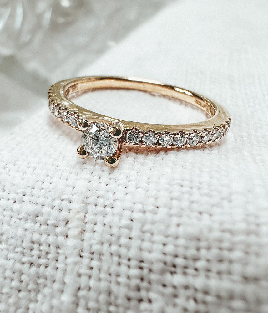 De Schitterende Geelgouden Solitair Ring met Zijstenen van Excellent Jewelry (14 karaat goud, 0.32ct diamant) schittert prachtig op een witte stoftextuur met briljant geslepen middensteen en H/SI-1 diamanten langs de band.