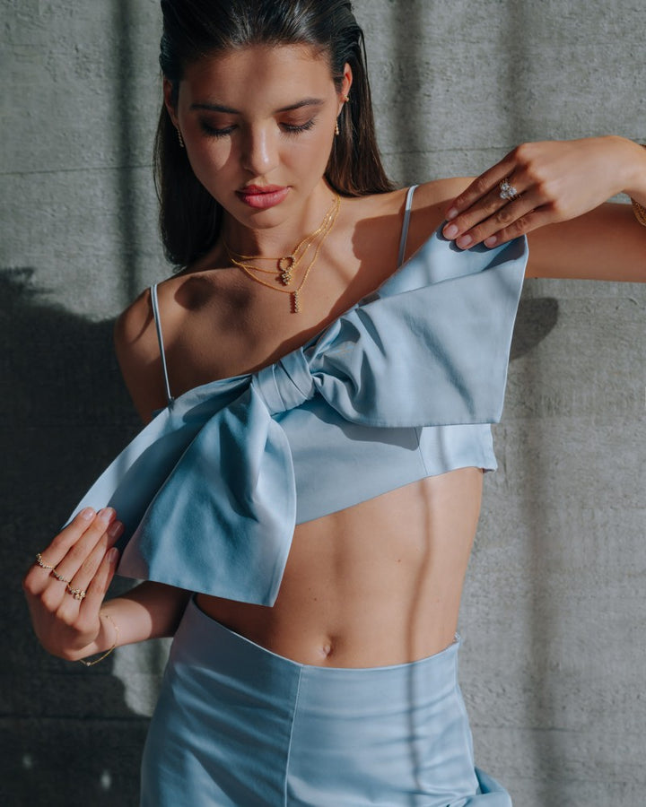 Een vrouw in een lichtblauwe, asymmetrische crop top en rok staat tegen een muur met textuur en kijkt naar beneden terwijl ze haar outfit aanpast. Ze draagt de Elegante 14k Geelgouden Ring met 0,81ct Diamanten uit de 'Bubbles' Collectie van Excellent Jewelry.
