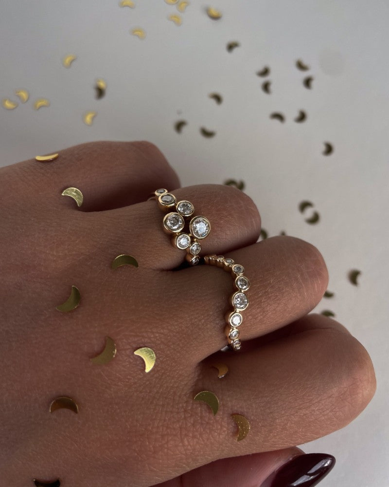 Een hand met gouden halvemaantjes confetti houdt een Elegante 14k Geelgouden Ring met 0,81ct diamanten uit de speelse 'Bubbles' Collectie van Excellent Jewelry vast. Ronde, fonkelende stenen schitteren terwijl meer halvemaanvormige confetti op de achtergrond strooit.