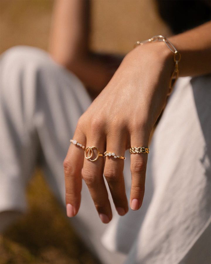 Een hand draagt meerdere dames sieraden, waaronder de Elegante Geelgouden Ring met Zirkonia's (14 karaat) van Excellent Jewelry, en een gouden armband. Zonlicht accentueert de sieraden terwijl de drager in witte broek buiten op de grond zit.