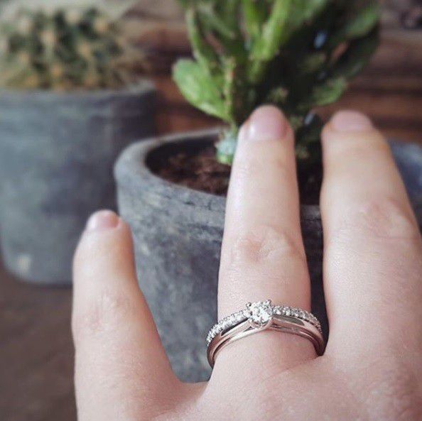 Een close-up van een hand die de Excellent Jewelry Witgouden Ring met Briljant Diamant - Subtiele Elegantie voor Elke Gelegenheid 0.25 crt. draagt, met een trouwring en twee vage potcactussen op de achtergrond.