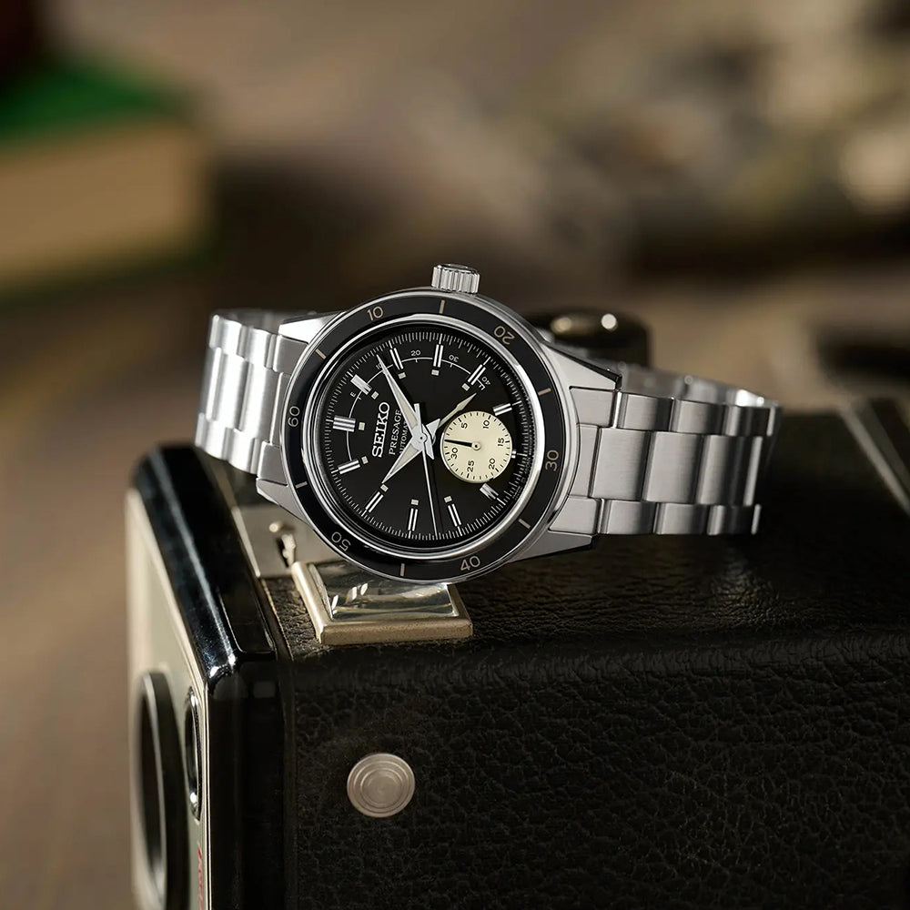 Het Seiko Presage heren horloge SSA449J1 rust elegant op een vintage camera, met een zwarte wijzerplaat en subdial. De strakke metalen band complementeert de zilveren, roestvrijstalen kast, terwijl een wazige boekachtergrond de tijdloze aantrekkingskracht versterkt.