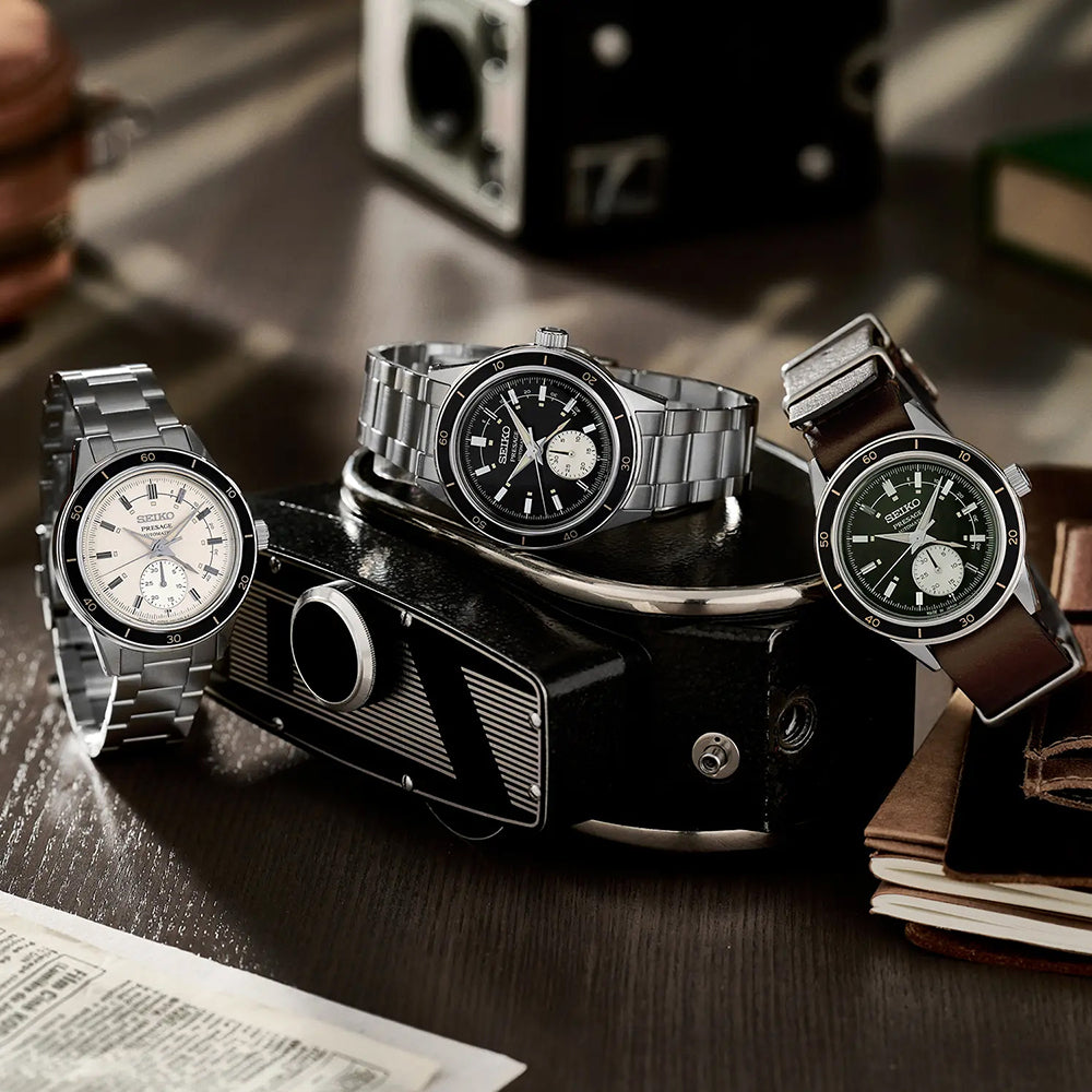 Drie polshorloges, waaronder een Seiko Presage SSA449J1 met een automatisch uurwerk in een roestvrijstalen kast, worden kunstig tentoongesteld op een vintage camera op een houten tafel. De horloges hebben witte, zwarte en groene wijzerplaten met metalen en leren banden. Boeken en papieren liggen nonchalant op de achtergrond.