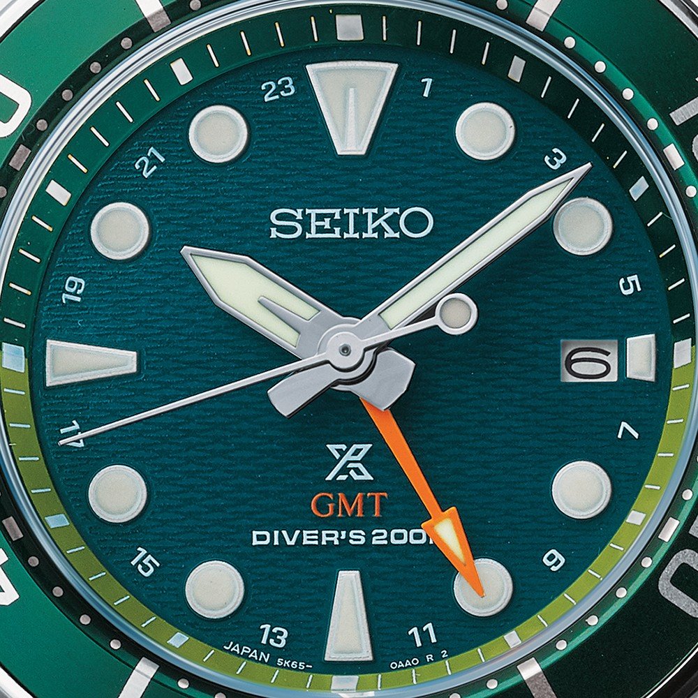 Een close-up van het Seiko Prospex heren horloge SFK003J1 toont de duikerswijzerplaat tegen een groene gestructureerde achtergrond. Het heeft lichtgevende markeringen en wijzers, een datumvenster op 3 uur, een oranje GMT-wijzer en is gemarkeerd met DIVERS 200 en JAPAN aan de onderkant.
