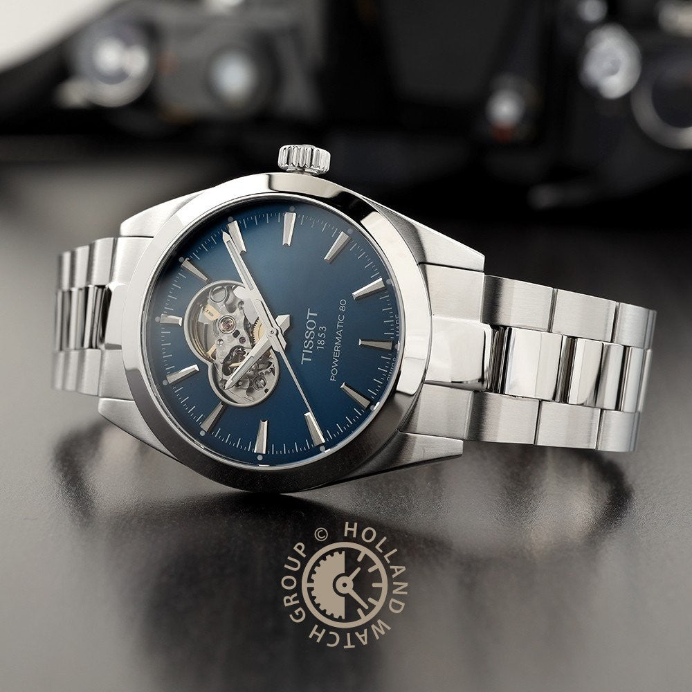 Het Tissot Gentleman Powermatic 80 Open Heart polshorloge, met een roestvrijstalen band en blauwe wijzerplaat, wordt getoond op een donker oppervlak met het Watch Group Holland logo eronder.