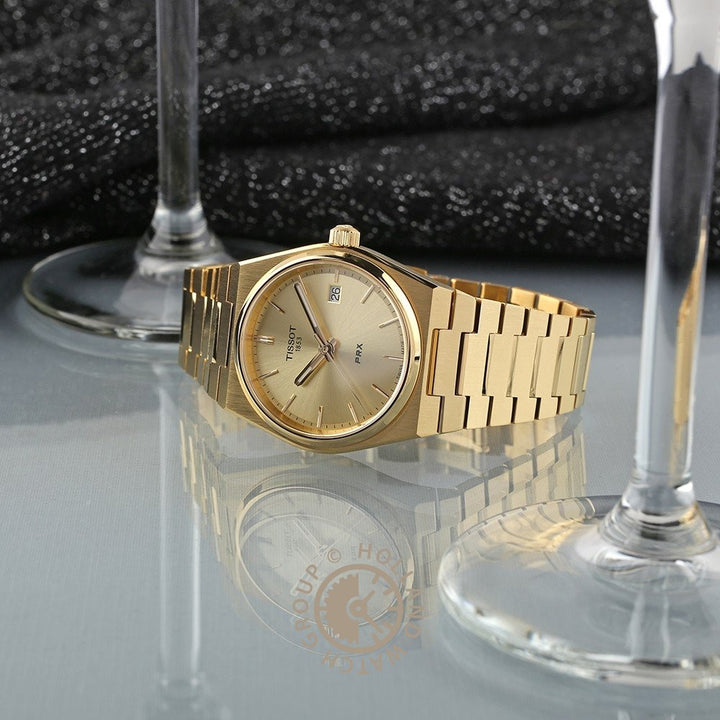 Een Tissot PRX 35mm gouden polshorloge met een metalen band wordt weergegeven op een reflecterend oppervlak tussen twee lege wijnglazen, tegen een donkere, glitterende stof. Dit Tissot horloge is voorzien van Zwitsers kwartsuurwerk en krasbestendig saffierglas.