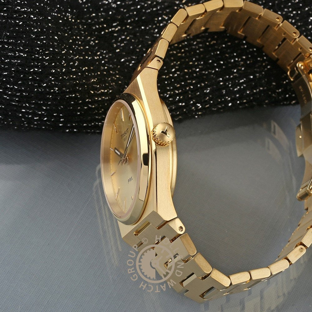 Het Tissot PRX polshorloge van 35 mm, met een gouden afwerking, Zwitsers kwartsuurwerk en een bijpassende metalen armband, wordt gepresenteerd op zwarte en grijze oppervlakken met textuur om het zijprofiel, de kroon en de gepolijste look te benadrukken.