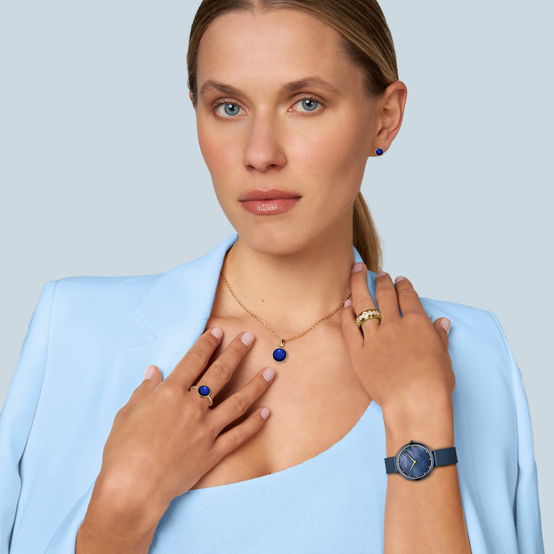 Een persoon in een lichtblauwe blazer en top draagt bijpassende blauwe en gouden sieraden, waaronder een ketting, ring, oorbellen en een elegant Bering dameshorloge (18132-399) met een Milanees-band. De achtergrond is effen lichtgrijs.